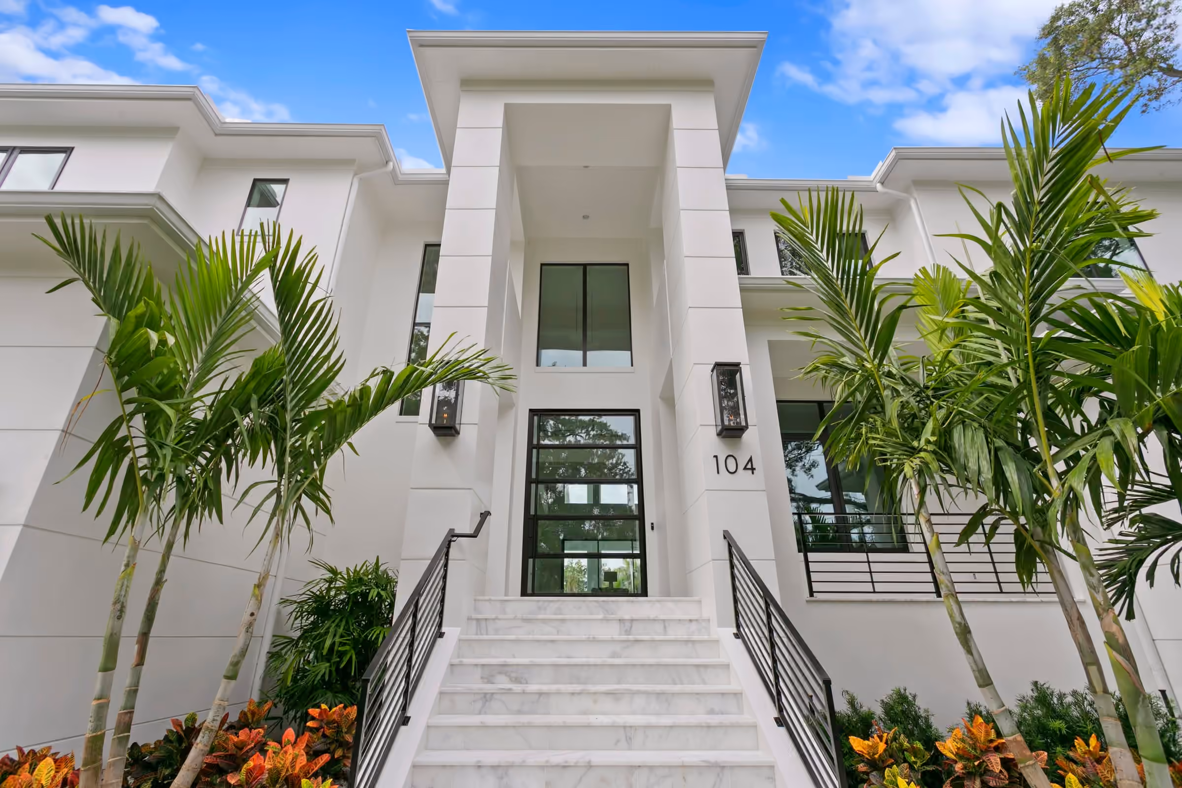 Front of a two-story white modern Halo home with wide stone steps, palms and a tall glass entry