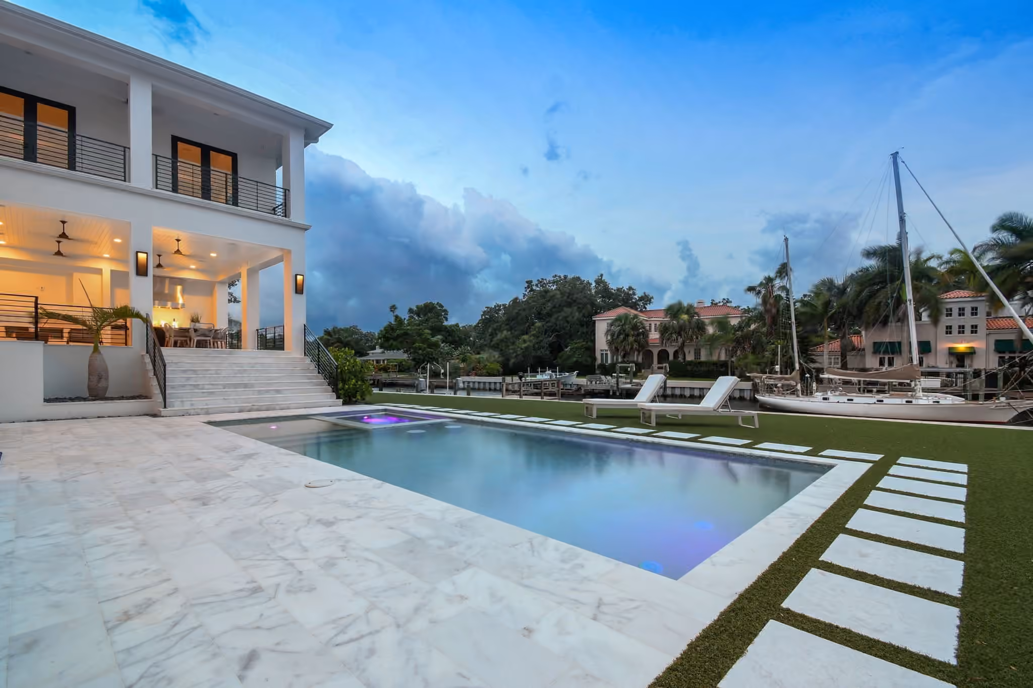 Rear of the home at dusk: lit covered porches, a marble pool deck and boats on the canal