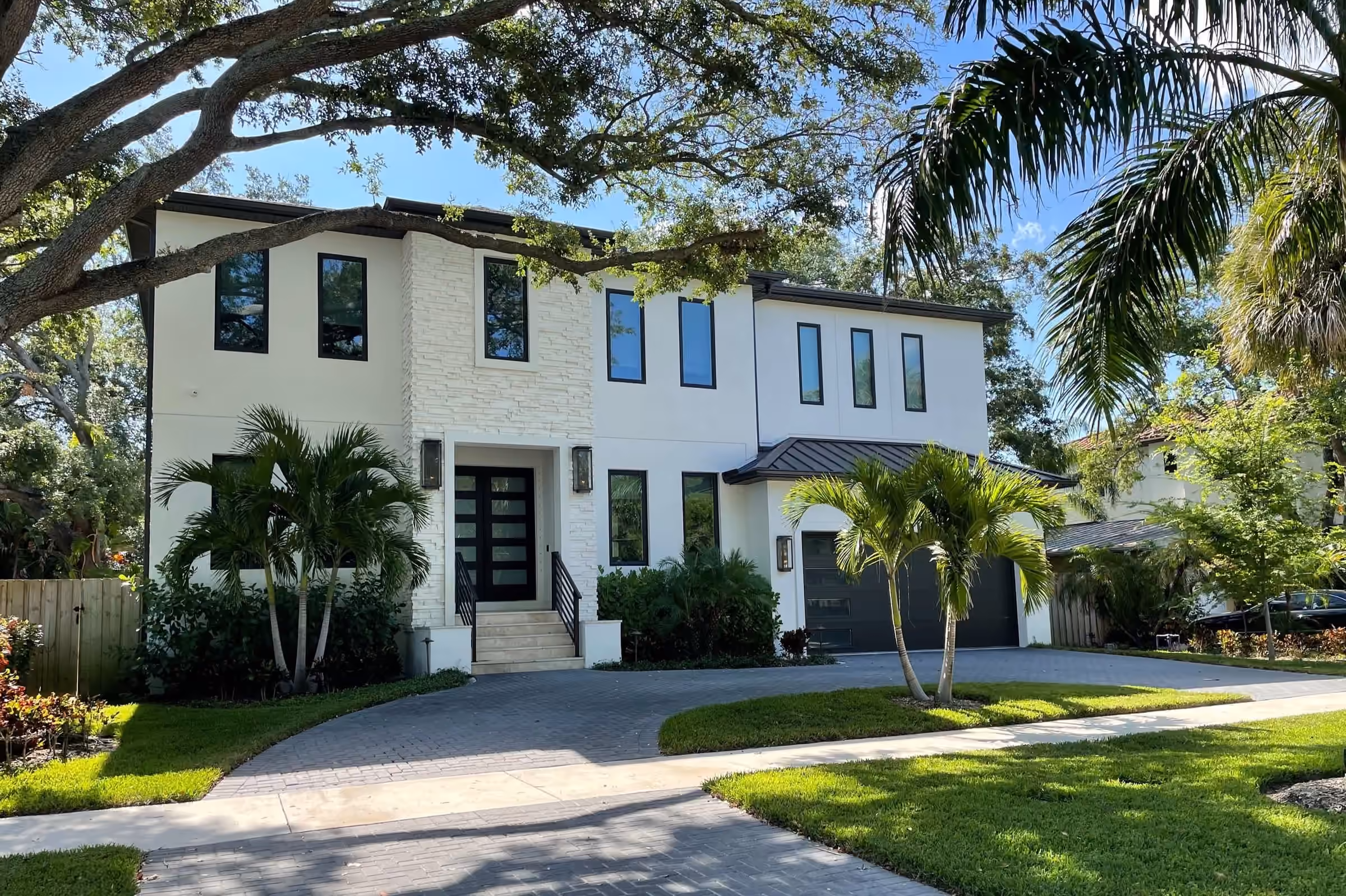 Two-story white modern home with black windows under oak trees and palms