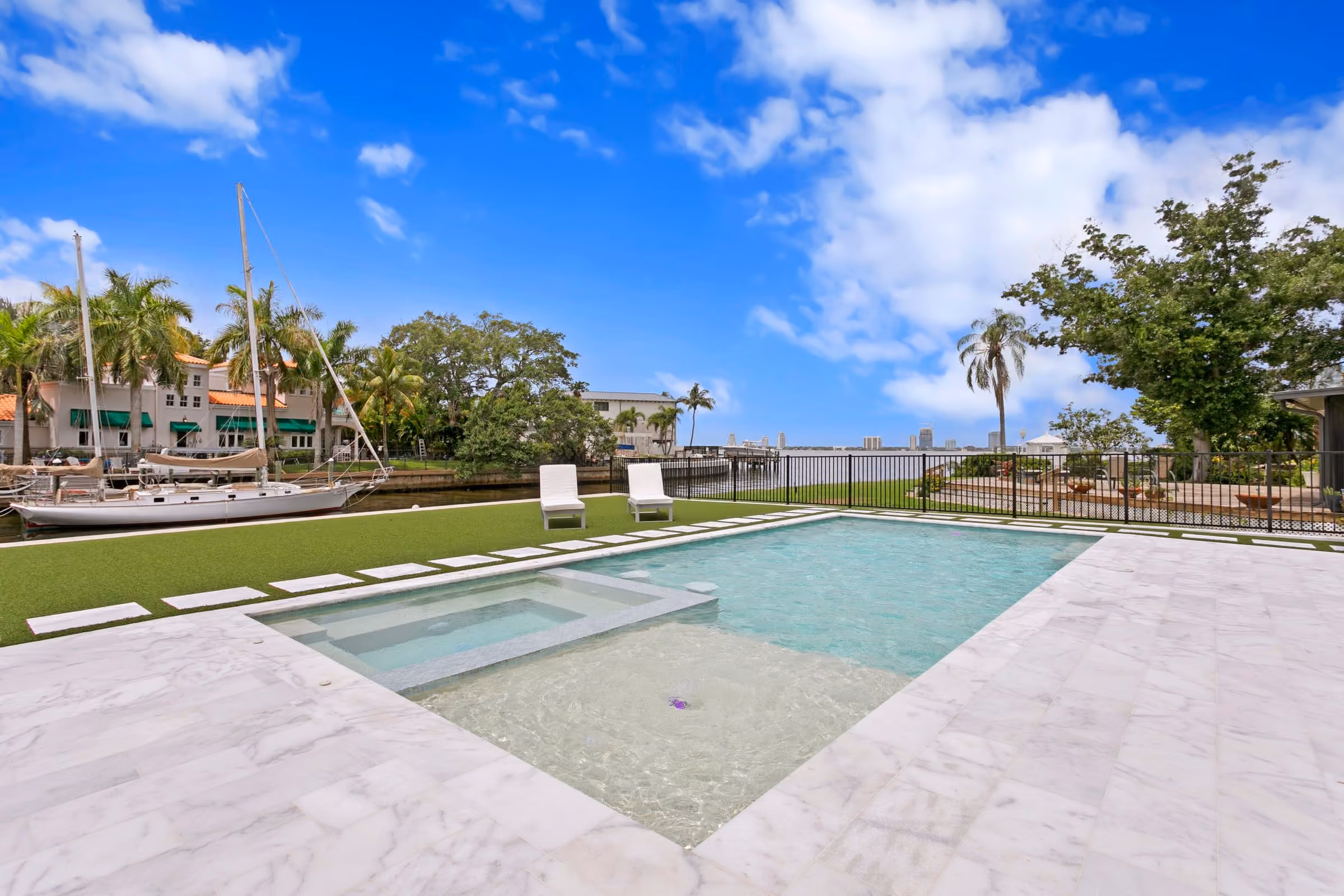 Pool and spa on a white stone deck by the water, with boat masts beyond